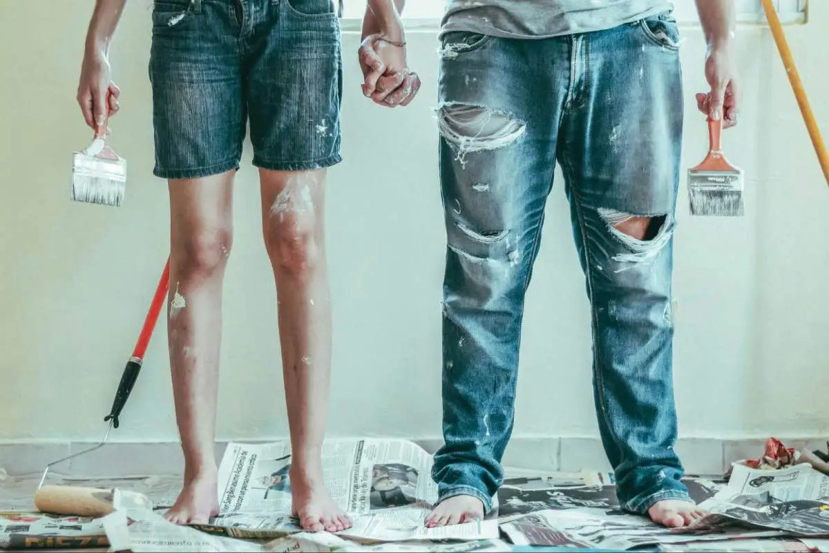 Two people from the waist down holding paint brushes and covered in paint on their pants and legs.